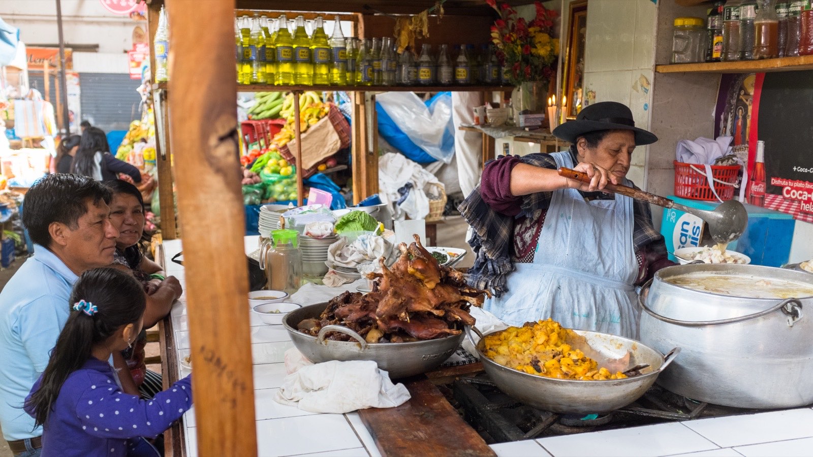 A cook ladling from a pot at a Cajamarca market food stall, trays of chicharron and mote in front of her