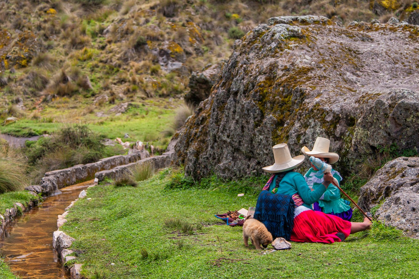 Two women in wide white hats sitting on the grass beside the open stone water channel at Cumbemayo